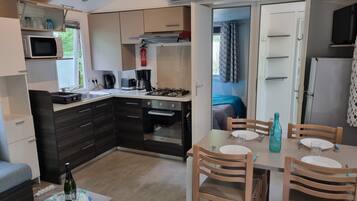A compact kitchen with modern appliances, a dining area with wooden chairs, and a visible bed in the adjacent room.