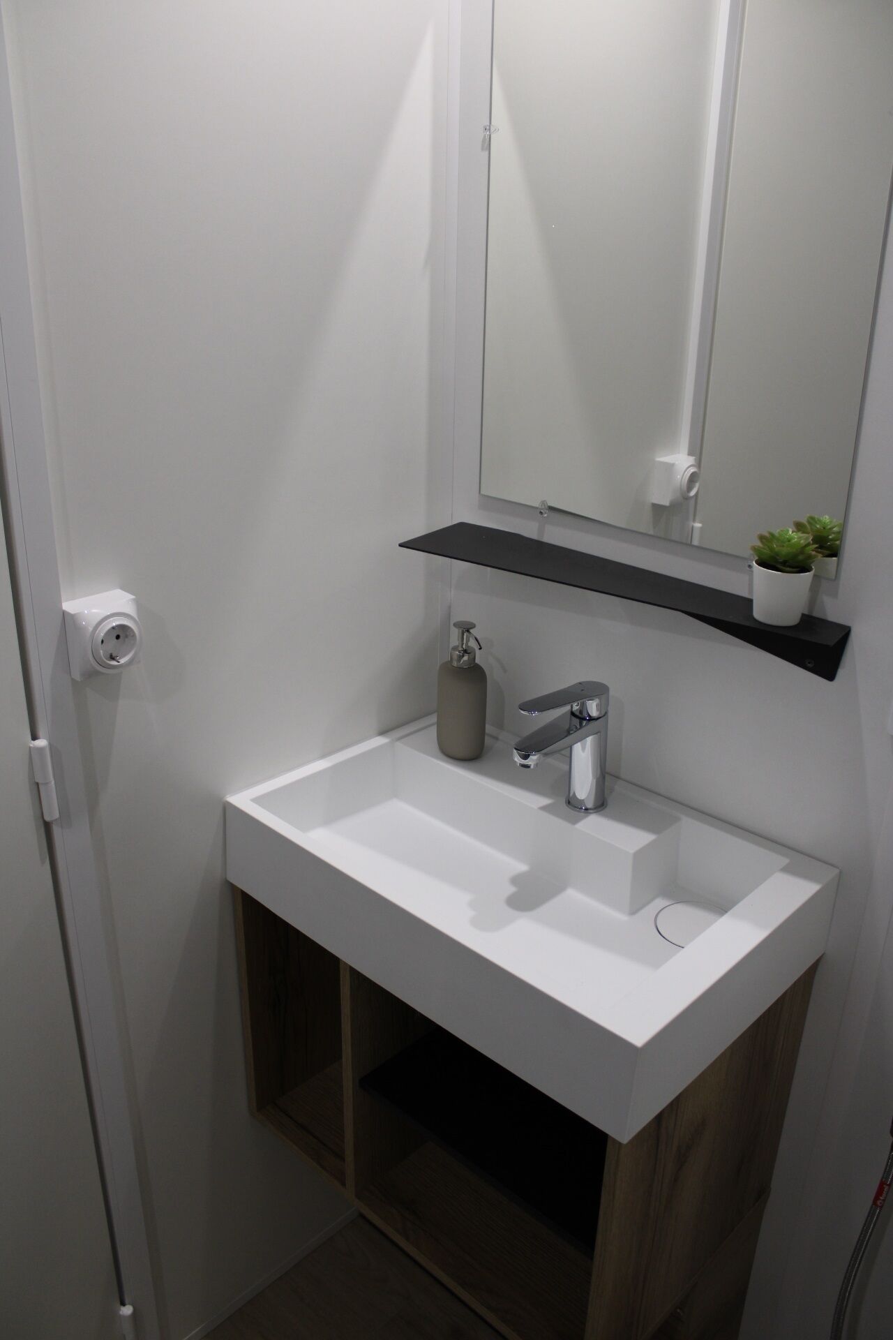 Un baño con un lavamanos blanco, un espejo, una repisa con una planta y un inodoro.