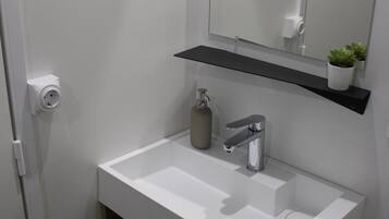 Un baño con un lavamanos blanco, un espejo, una repisa con una planta y un inodoro.