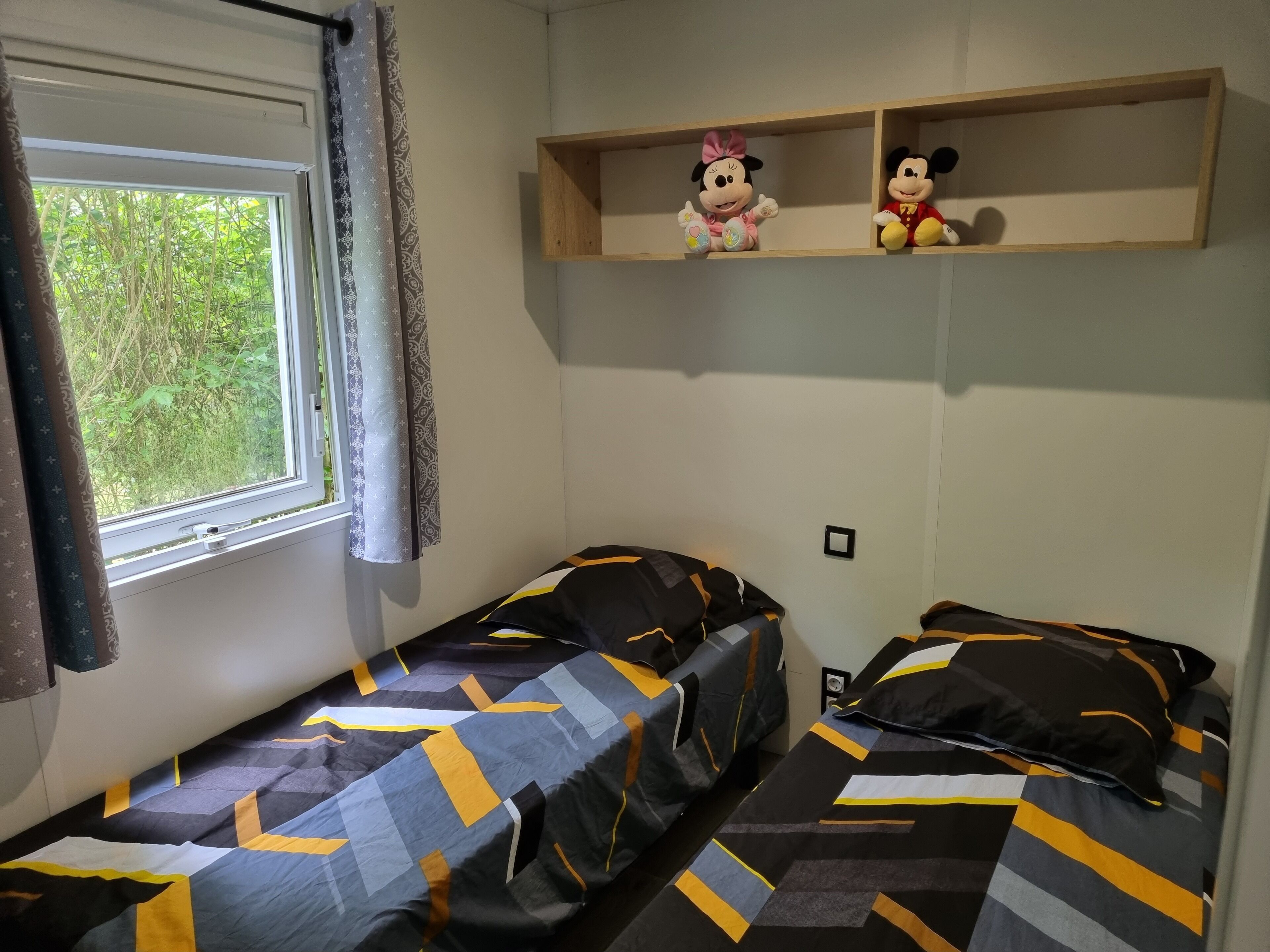 Una habitación con dos camas, una ventana con cortinas y dos estantes con peluches.