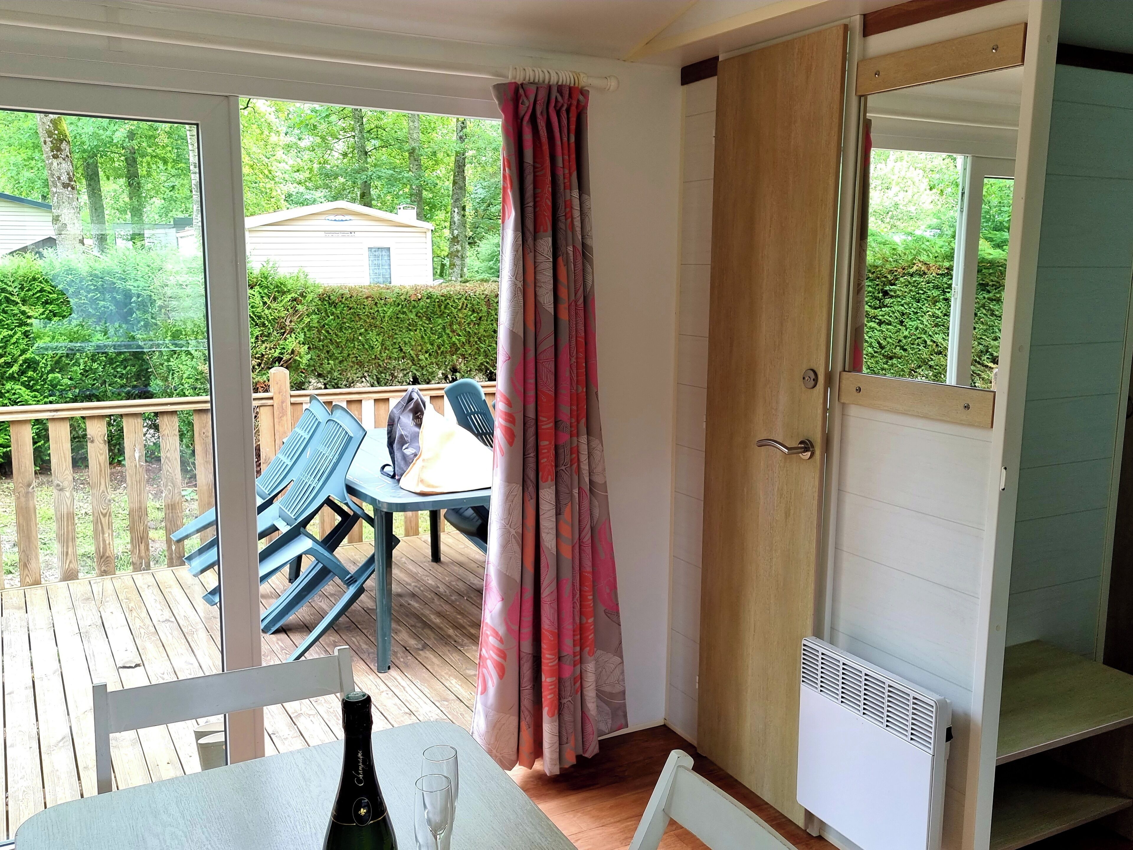 Ein Zimmer mit einer Holzterrasse, einem Tisch mit Flasche und Gläsern sowie Blick auf ein Haus und Grünfläche.