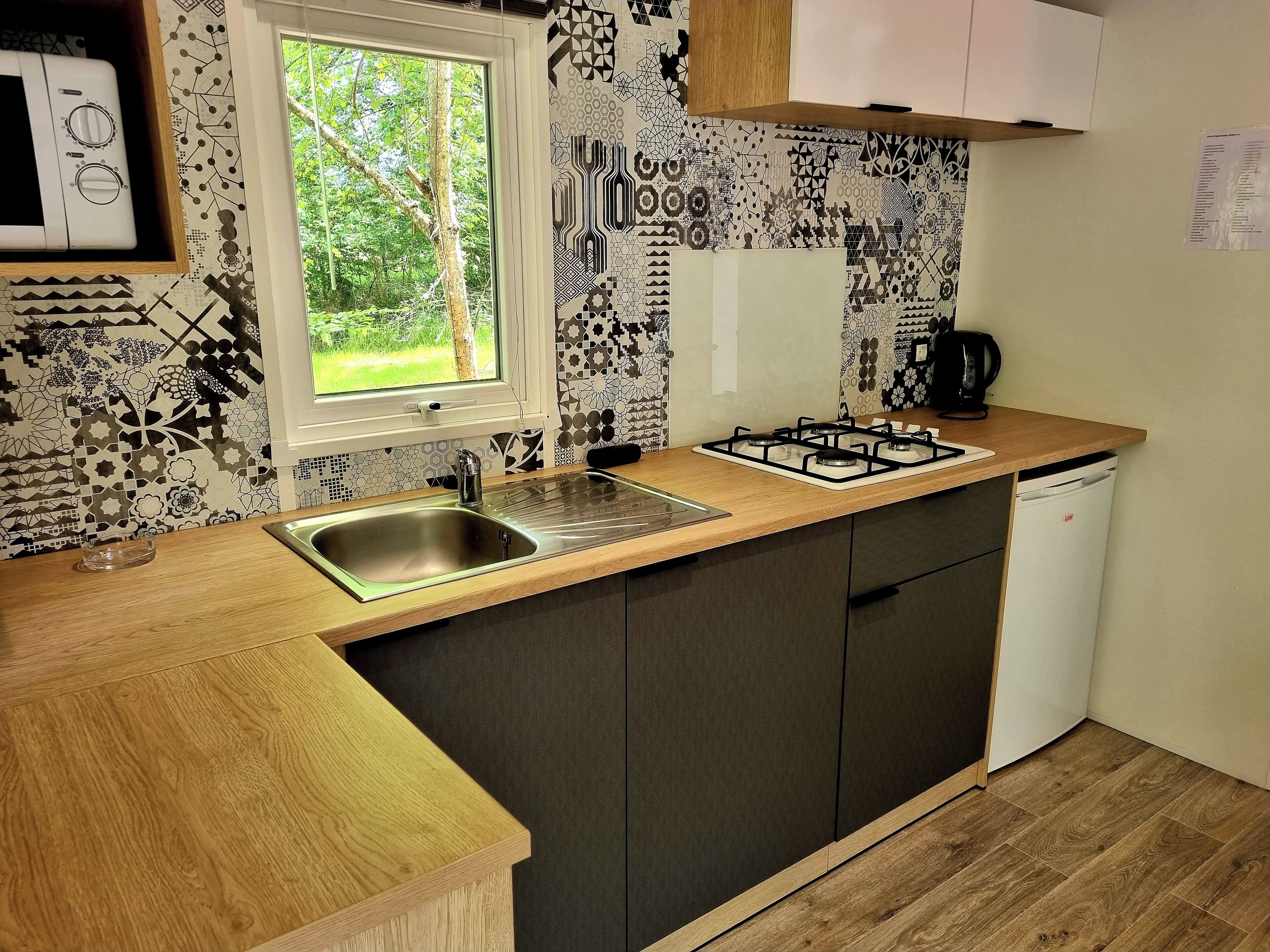 A compact kitchen with a sink, stove, and white dishwasher. A window provides a view of greenery outside.