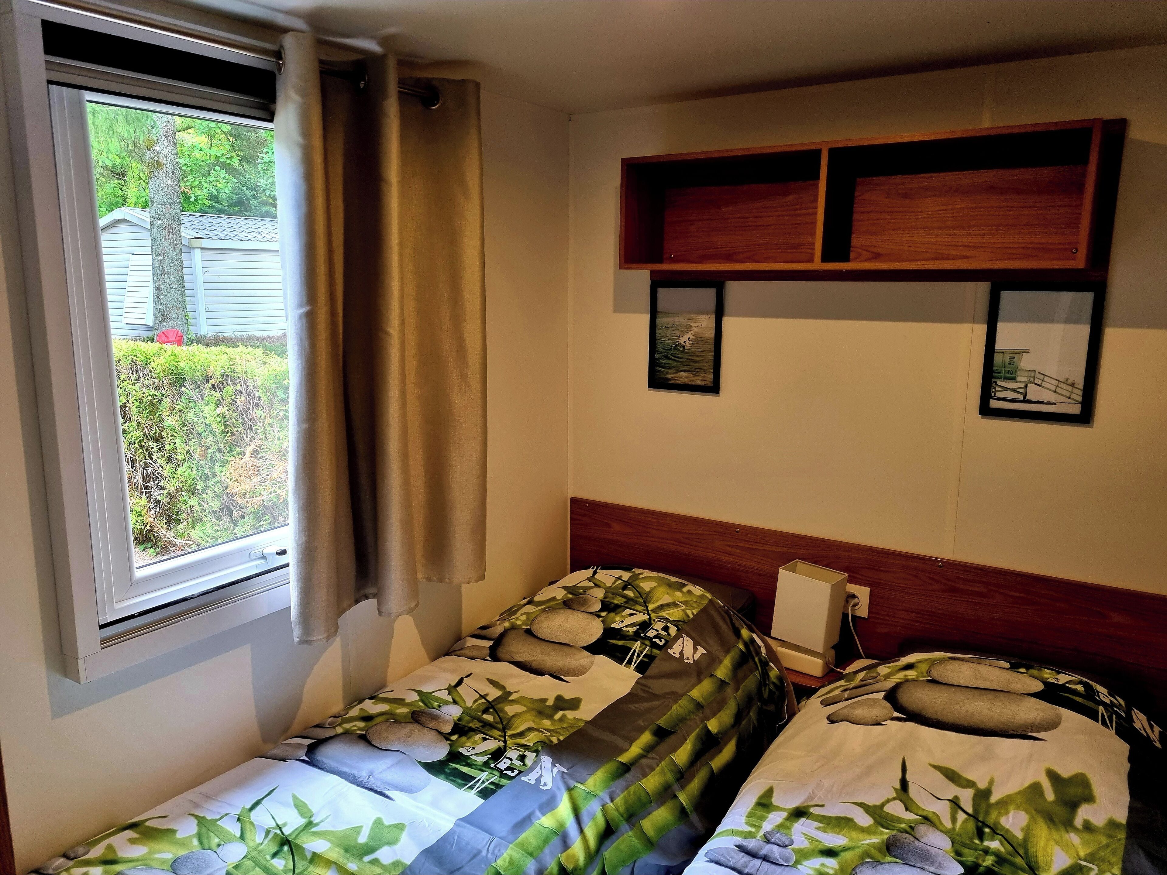 A bedroom with two single beds, a window with curtains, a wooden headboard, and a small shelf with pictures.