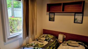 A bedroom with two single beds, a window with curtains, a wooden headboard, and a small shelf with pictures.