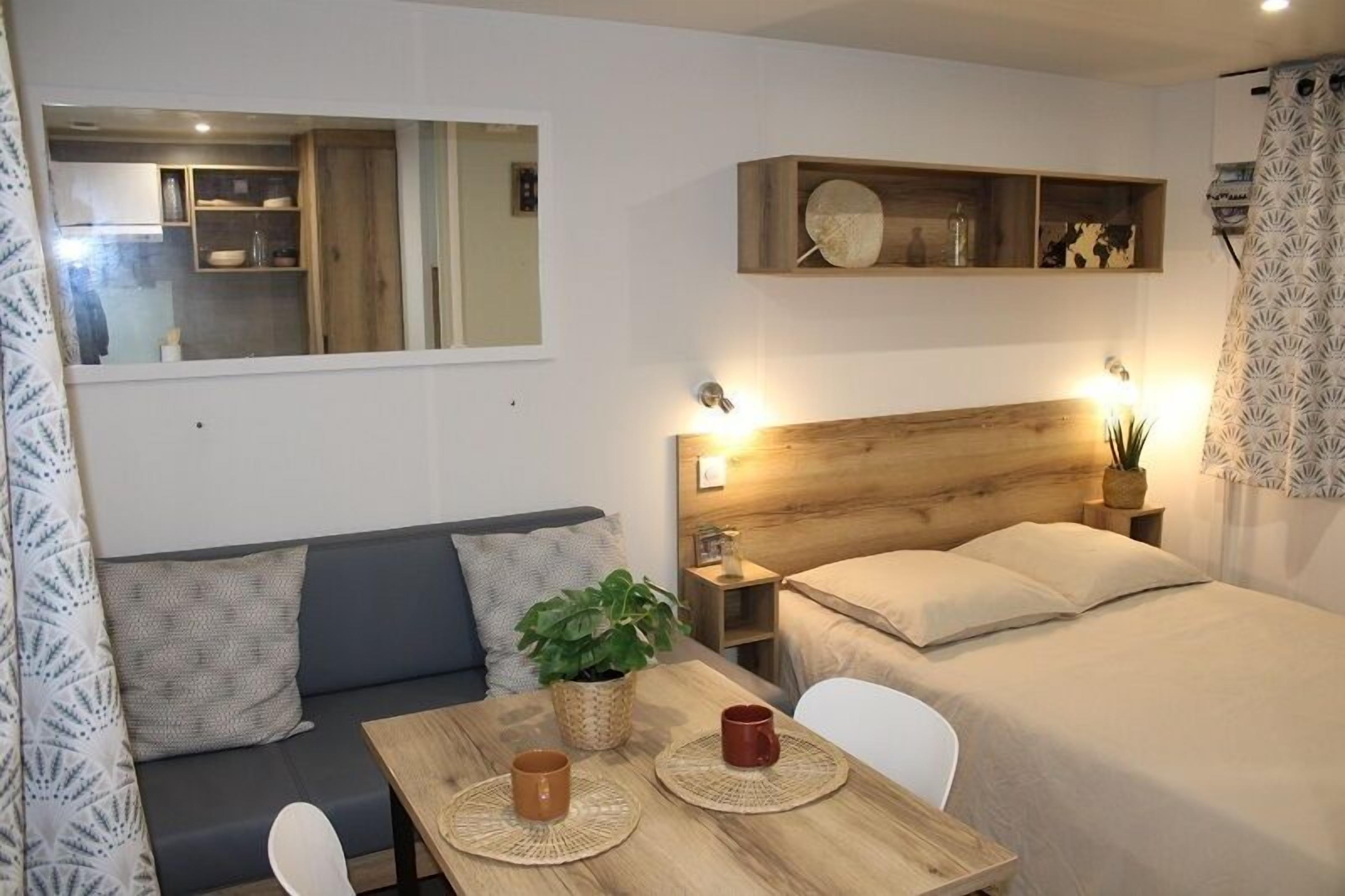 Une pièce compacte et bien éclairée, comprenant un lit, une petite table avec deux tasses, un canapé, une tête de lit en bois et un miroir qui reflète un coin cuisine.
