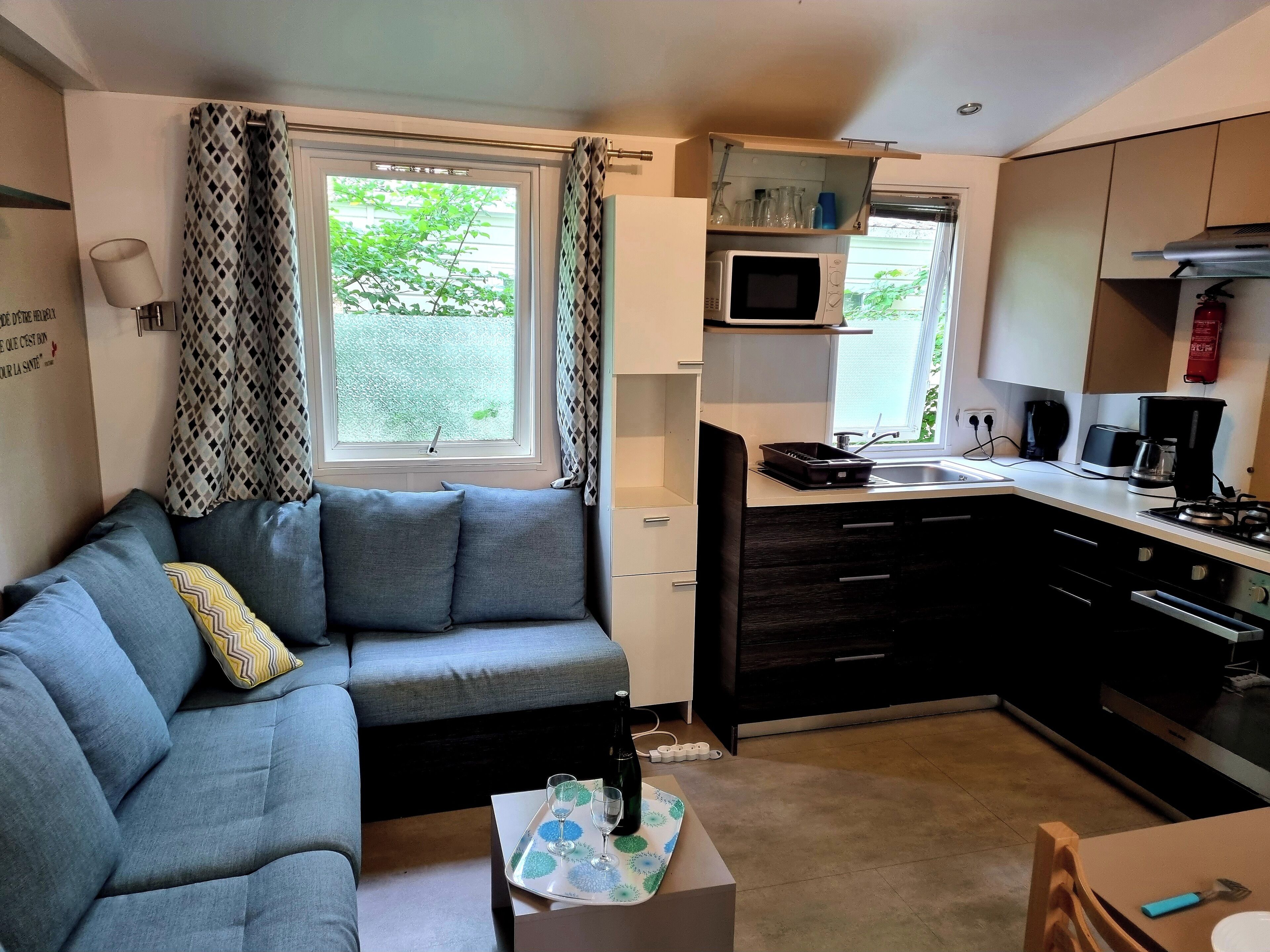 A compact living space with a sofa, a small table with a bottle and glasses, a kitchenette with a microwave, stove, and cabinets, and a window with curtains.