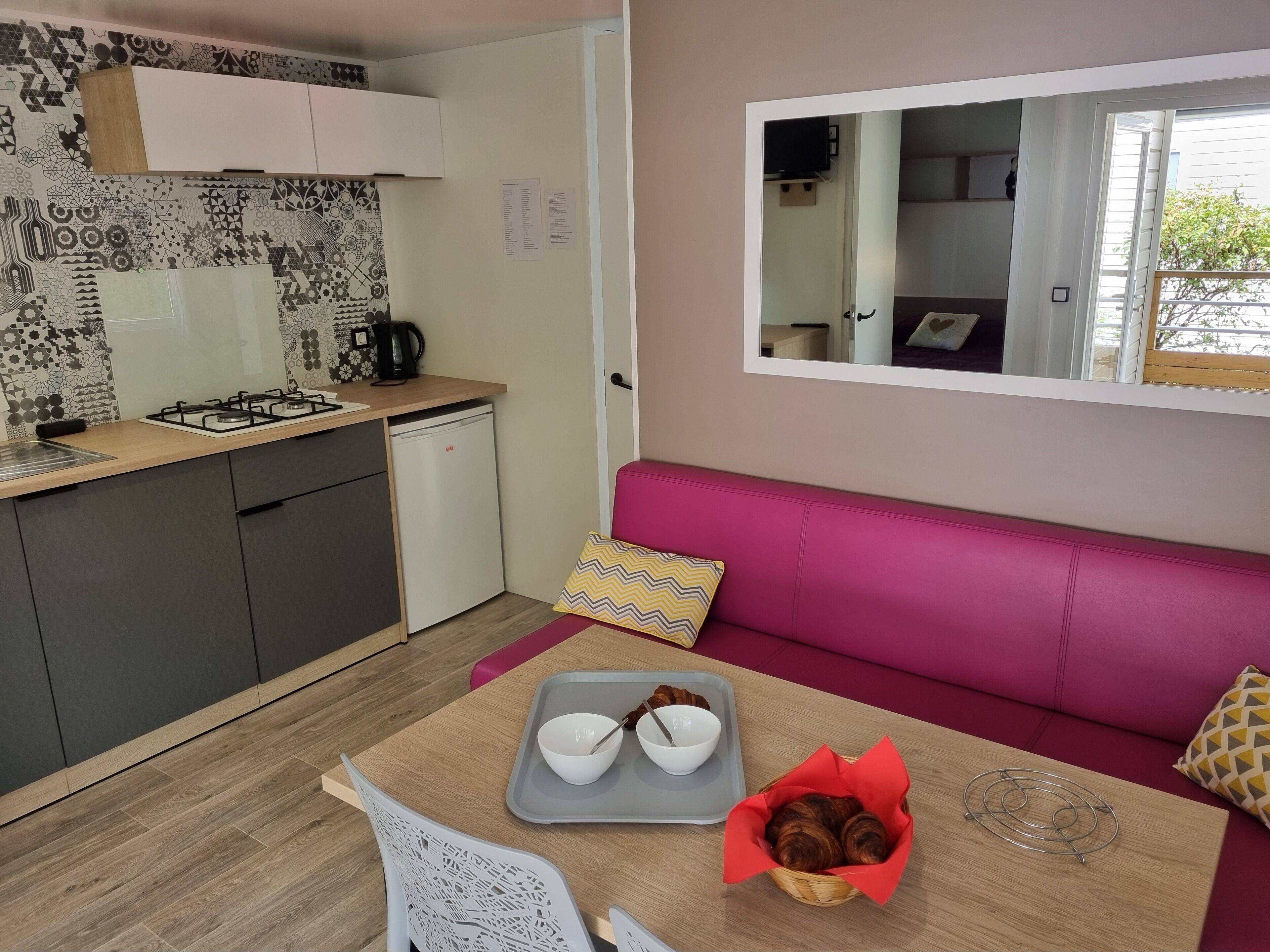 Cocina compacta con estufa a gas, refrigerador pequeño y azulejos decorativos en el salpicadero. Se ve un sofá rosa con un espejo arriba, y una mesa de comedor con una bandeja de croissants y tazones al frente.