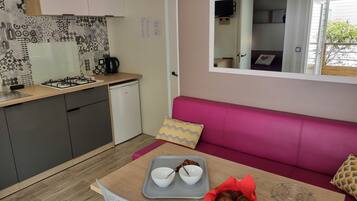 Cocina compacta con estufa a gas, refrigerador pequeño y azulejos decorativos en el salpicadero. Se ve un sofá rosa con un espejo arriba, y una mesa de comedor con una bandeja de croissants y tazones al frente.