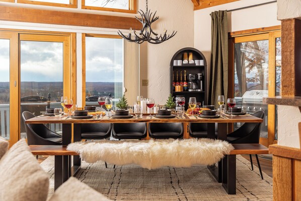 Un comedor con una mesa grande dispuesta para seis personas, una araña y vistas del paisaje a través de amplios ventanales.