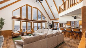 A spacious living area with a large sofa, wooden flooring, and a view through large windows. - Amazing Views! Jacuzzi-Sauna-Firepit-Arcades-Game Room-King Beds-Walk to Schuss (Bellaire)
