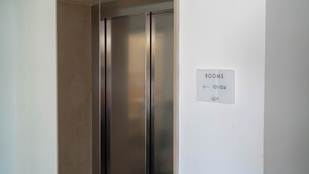 Un ascensor moderno con revestimiento metálico, un cartel que indica “HABITACIONES 101-104” y una vista parcial de una pared color beige.