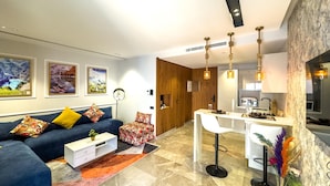 Una sala moderna con un sofá azul, cojines coloridos, una mesa de centro blanca y una zona de cocina con una barra blanca y taburetes.