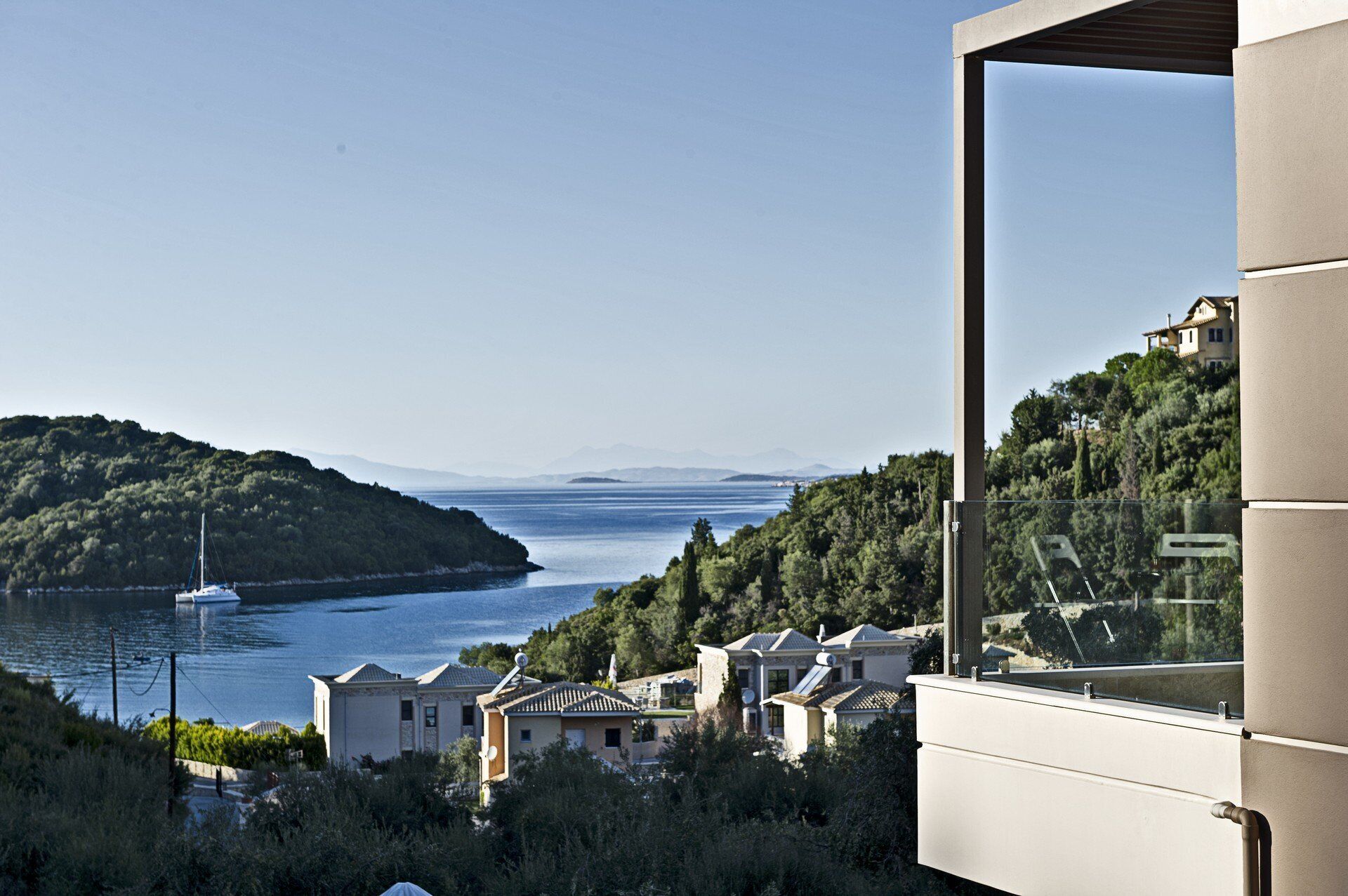 A coastal view with a sailboat on the water, houses on a hillside, and a modern building with a large window.