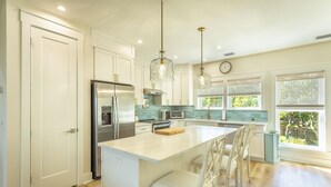 Une cuisine moderne avec des meubles blancs, un réfrigérateur en acier inoxydable, un îlot central intégré avec des sièges, des luminaires suspendus et une vue sur l’extérieur grâce à de grandes fenêtres.