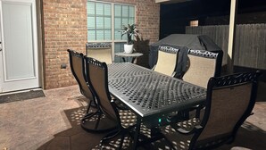 En uteplats med ett metallbord och stolar, en överbyggd grill och en mur av tegelsten.