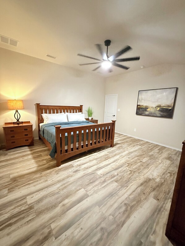 A bedroom with a wooden bed, a nightstand, a lamp, a ceiling fan, and a painting on the wall.