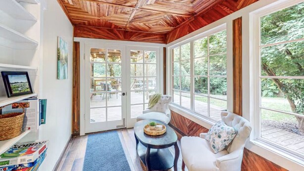 A cozy room with a wooden ceiling, large windows, a white sofa, a small round table, and a bookshelf. - Lucky Clover over the Covered Bridge (Eureka)