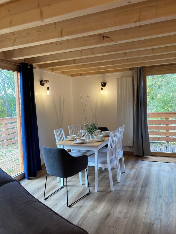 Comedor con mesa de madera dispuesta para cuatro personas, rodeada de sillas blancas. Hay dos lámparas fijadas a la pared, un ventanal con vista a la vegetación y una puerta de madera grande.