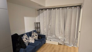 Una sala de estar con un sofá azul, cojines estampados, un bolso negro y una cortina blanca.