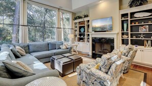 Una sala de estar moderna con un sofá gris, sillones estampados, una chimenea y un ventanal grande con vistas a árboles.