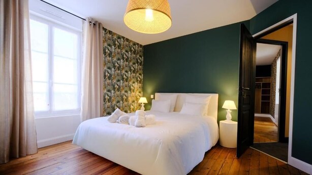 A neatly made bed with white linens and towels, a large window with curtains, a patterned wall, and a door leading to another room. - LOGIS DES COEVRONS (Évron)
