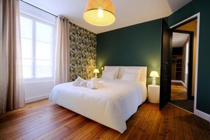Una cama bien tendida con sábanas y toallas blancas, un ventanal con cortinas, un papel pintado y una puerta que da a otra habitación.