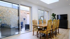 A dining area with a wooden table and floral chairs, a television, and a stone wall visible through glass doors. - LOGIS DES COEVRONS (Évron)
