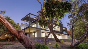 Une maison moderne à deux étages, dotée d’un balcon et entourée d’arbres et de plantes.