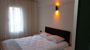 A neatly made bed with a white duvet and two patterned pillows, a headboard with a light fixture, and a window with sheer curtains. - Rayon Apart hotel (Marmaris)