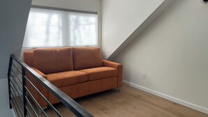 Une pièce avec un plancher en bois, un canapé marron, une rambarde noire et une fenêtre avec des stores.