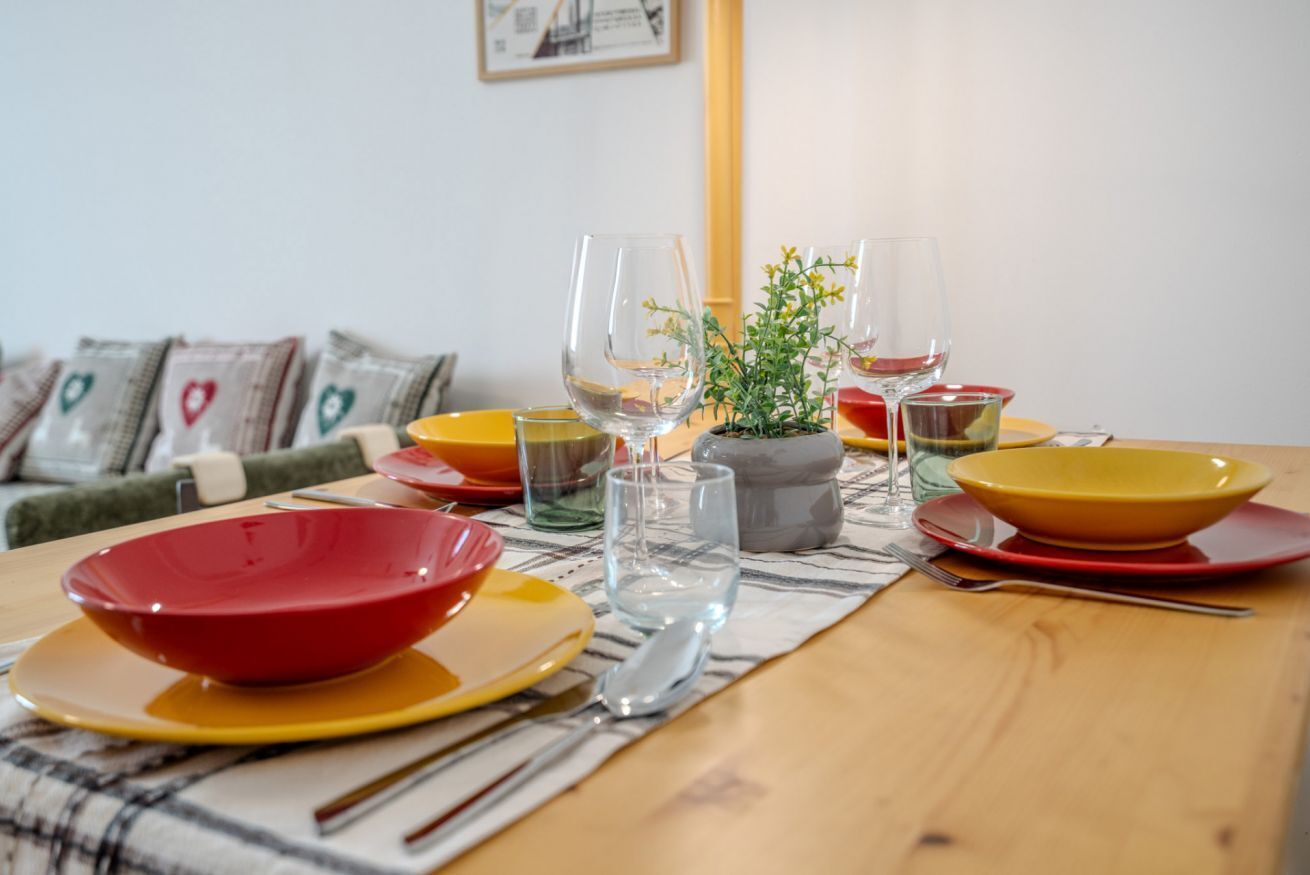 Une table à manger dressée avec des bols, des verres et des couverts aux couleurs vives, agrémentée d’une plante en guise de centre de table et d’un tableau encadré accroché au mur.