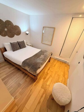 A bedroom with a bed, wooden flooring, a white wall, a ceiling fan, and a small white stool. - 403 | Luxury Condo | Incredible View & Pool (Baie-Saint-Paul)