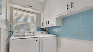 A laundry room with two white washers and dryers, white cabinets, and blue-tiled walls. - 2589 Bayshore Dr (Matlacha)