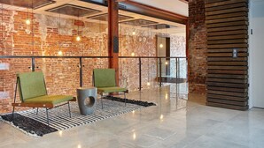 Um hall moderno com paredes de tijolo aparente, piso de concreto polido e diversos assentos, incluindo uma poltrona estofada verde e um banquinho de metal.