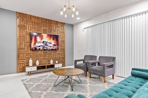 A modern living room with a flat-screen TV mounted on a wooden wall, two grey armchairs, a teal sofa, a round coffee table, and a patterned rug. - 5BR townhouse sleeps 13 with private pool (Kissimmee)