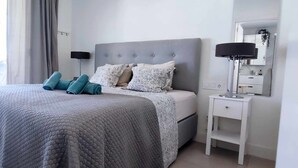A neatly made bed with a grey quilt, two pillows, and a folded towel. There is a bedside table with a lamp and a small shelf. - Panorama Beach - Penthouse (Torrox)
