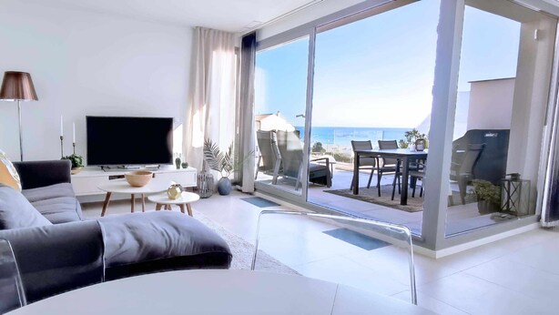 A modern living room with a large flat-screen TV, a grey sofa, a small coffee table, and a balcony with outdoor seating. - Panorama Beach - Penthouse (Torrox)