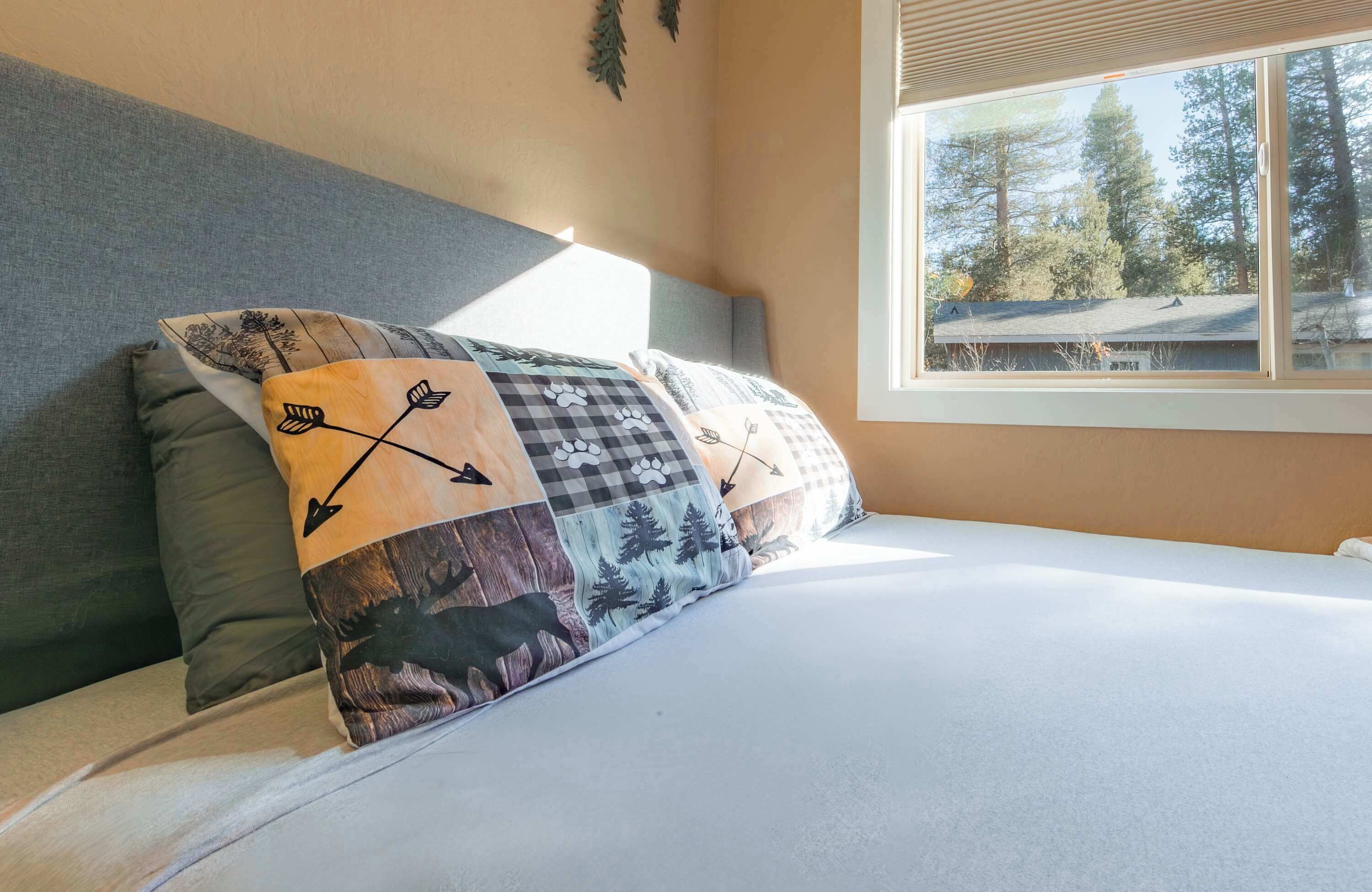 Een bed met meerdere kussens, een raam met uitzicht op bomen, en een wand met een plant.