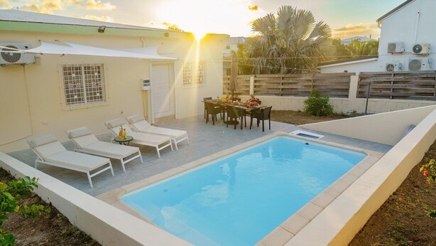 A swimming pool with lounge chairs and a table, surrounded by a low wall and a wooden fence. - Casa Verde: Grand Case Village (Grand-Case)
