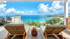 Un balcon agrémenté de deux fauteuils en bois et d’un vase décoratif, offrant une vue dégagée sur une mer bleue et un paysage montagneux.