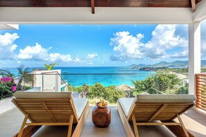 Ein Balkon mit zwei Holzliegen und einer dekorativen Vase, mit Blick auf das klare, blaue Meer und eine gebirgige Landschaft.