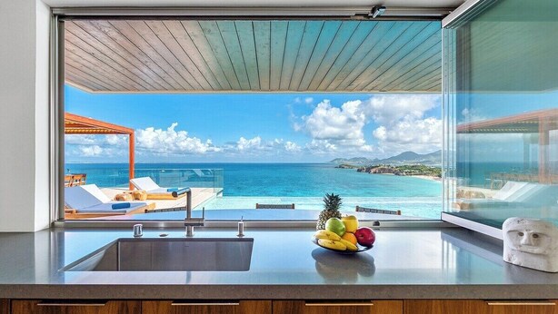 Une cuisine avec vue sur une piscine et une plage, un panier de fruits sur le plan de travail et des meubles de rangement en bois.