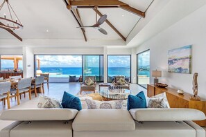 Ein großzügiges Wohnzimmer mit einem großen Fenster mit Meerblick, einem Essbereich mit Holzmöbeln und einem modernen Deckenventilator.