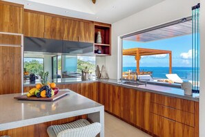 Eine moderne Küche mit Holzausstattung, einer großen Kochinsel mit Obst und Meerblick durch eine Glastür.