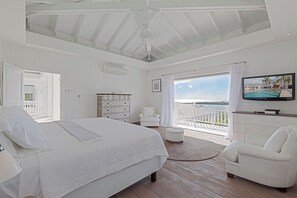Ein geräumiges Schlafzimmer mit einem großen Bett, einem Fernseher, einer weißen Kommode und einem Balkon mit Meerblick.