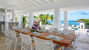 Un comedor con una mesa de madera dispuesta para seis personas, sillas blancas y vista al océano.