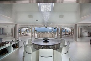 A spacious, modern interior with a central round table and white chairs, large windows offering a view of the ocean, and a high ceiling with a hanging light fixture. - C'est La Vie   Luxury beachfront villa (Saint Martin)