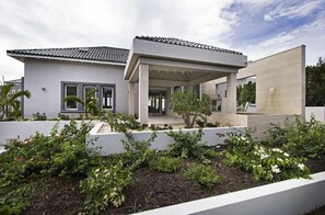 Une maison moderne à deux étages, dotée d’un patio couvert et entourée d’un jardin aménagé avec diverses plantes et arbustes.