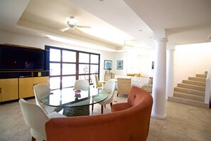 Ein geräumiges Wohnzimmer mit Esstisch aus Glas, bequemen Sitzmöglichkeiten, einem Flachbildfernseher und einer Treppe, die zu einer weiteren Ebene führt.