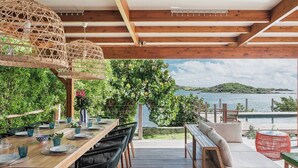 Ein Außenbereich zum Essen mit Holztischen und -stühlen, einer hängenden Lampenschirm aus Weidengeflecht und Blick auf das Meer.